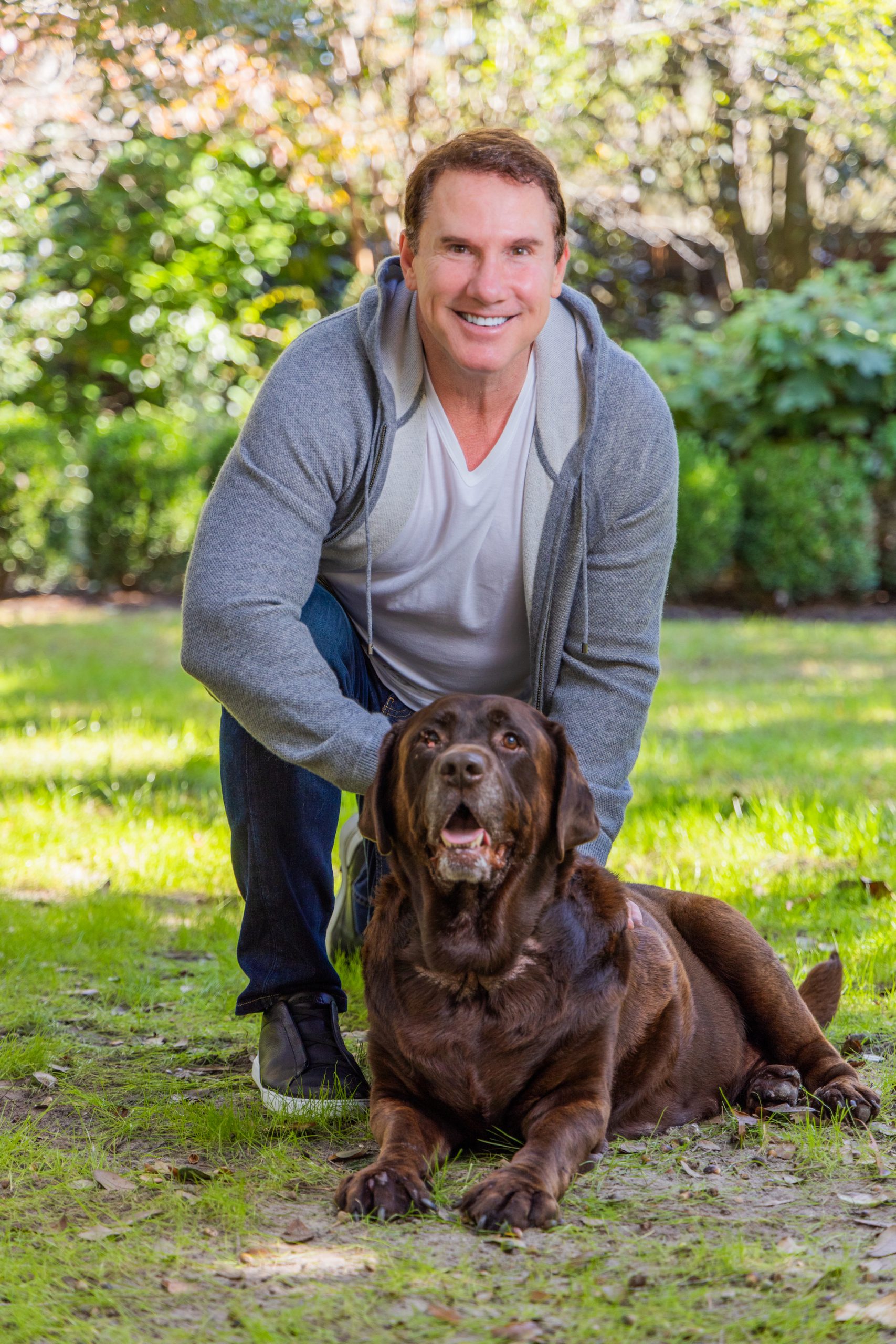 Smiling man in grey sweatshirt takes a knee and holds large brown dog that is lying down on a green lawn.